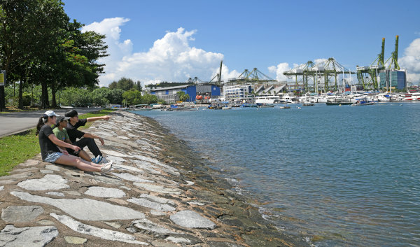 Three people sitting on a rocky embankment by the sea, looking at boats and harbor cranes.